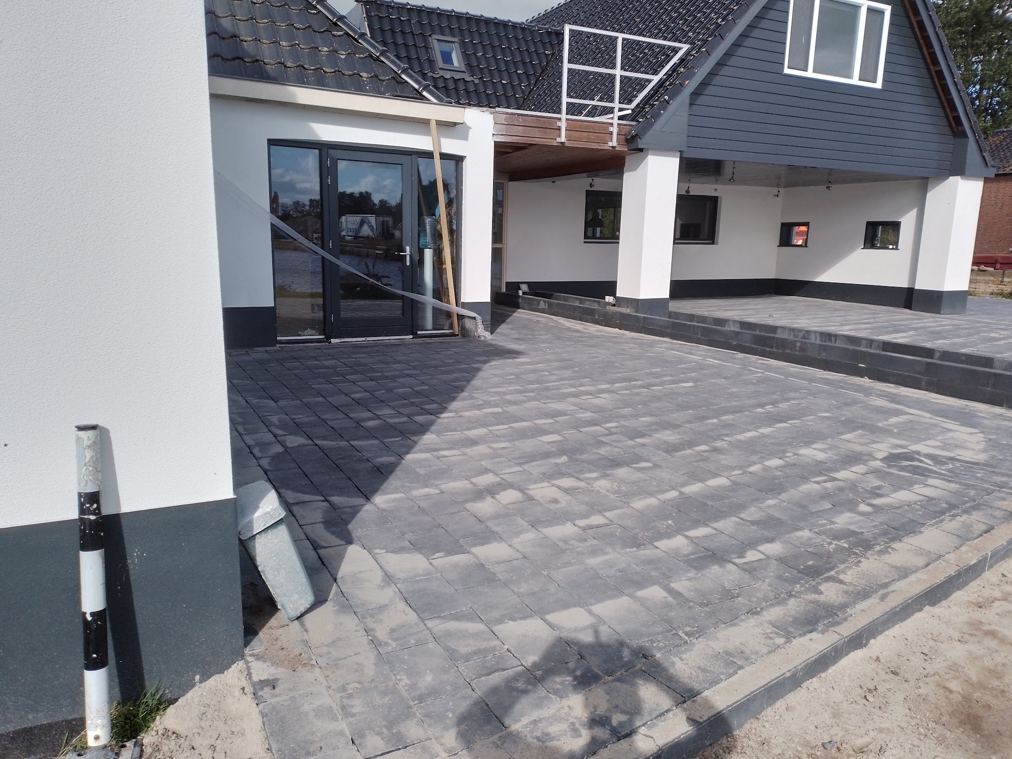 Antraciet bestraat erf voor een wit gestucte, verbouwde boerderij, met een traptrede naar de overkapping
