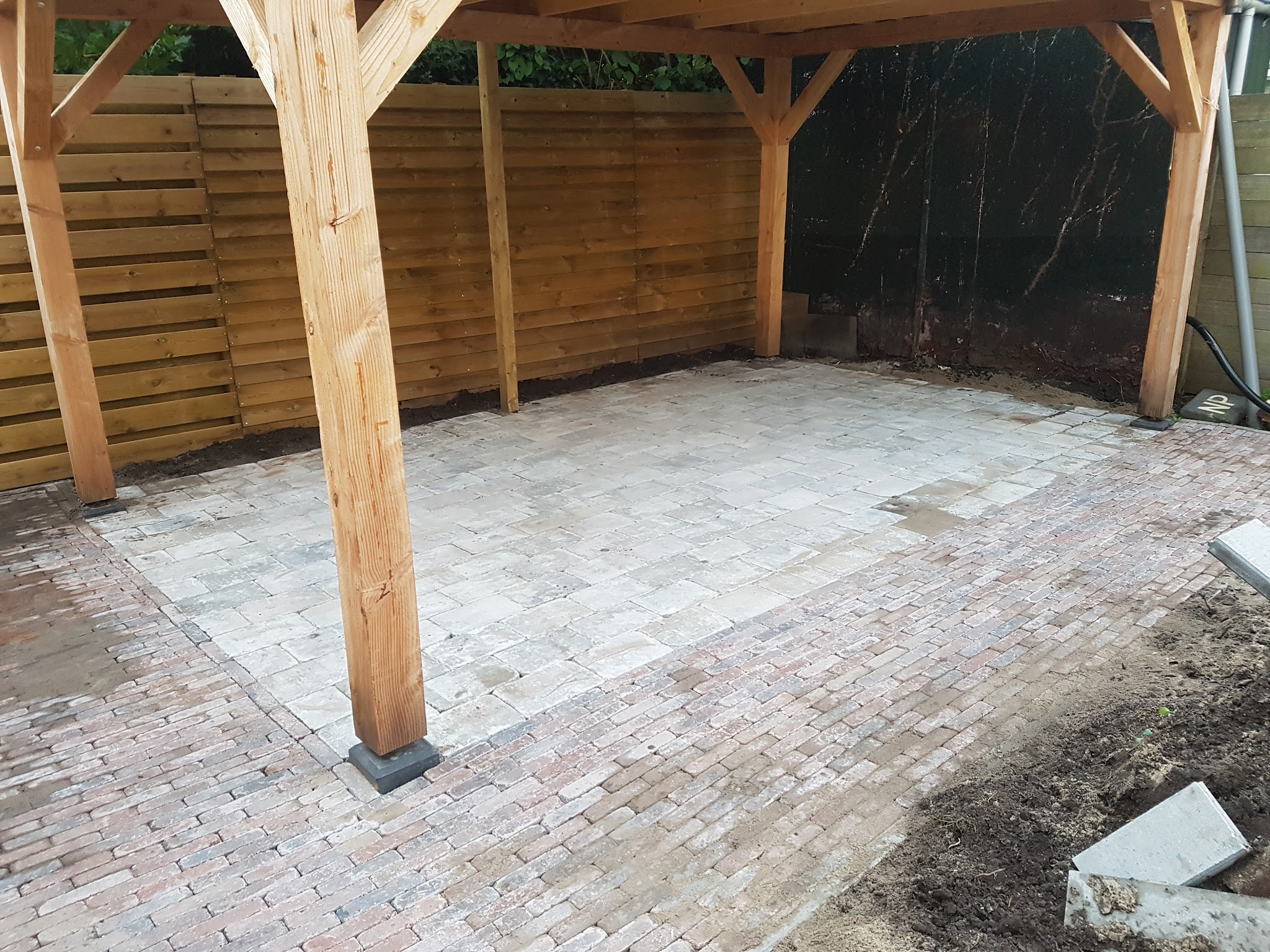Houten carport met een vloer van getrommelde lichte steen omrand door oude rode gebakken klinkers