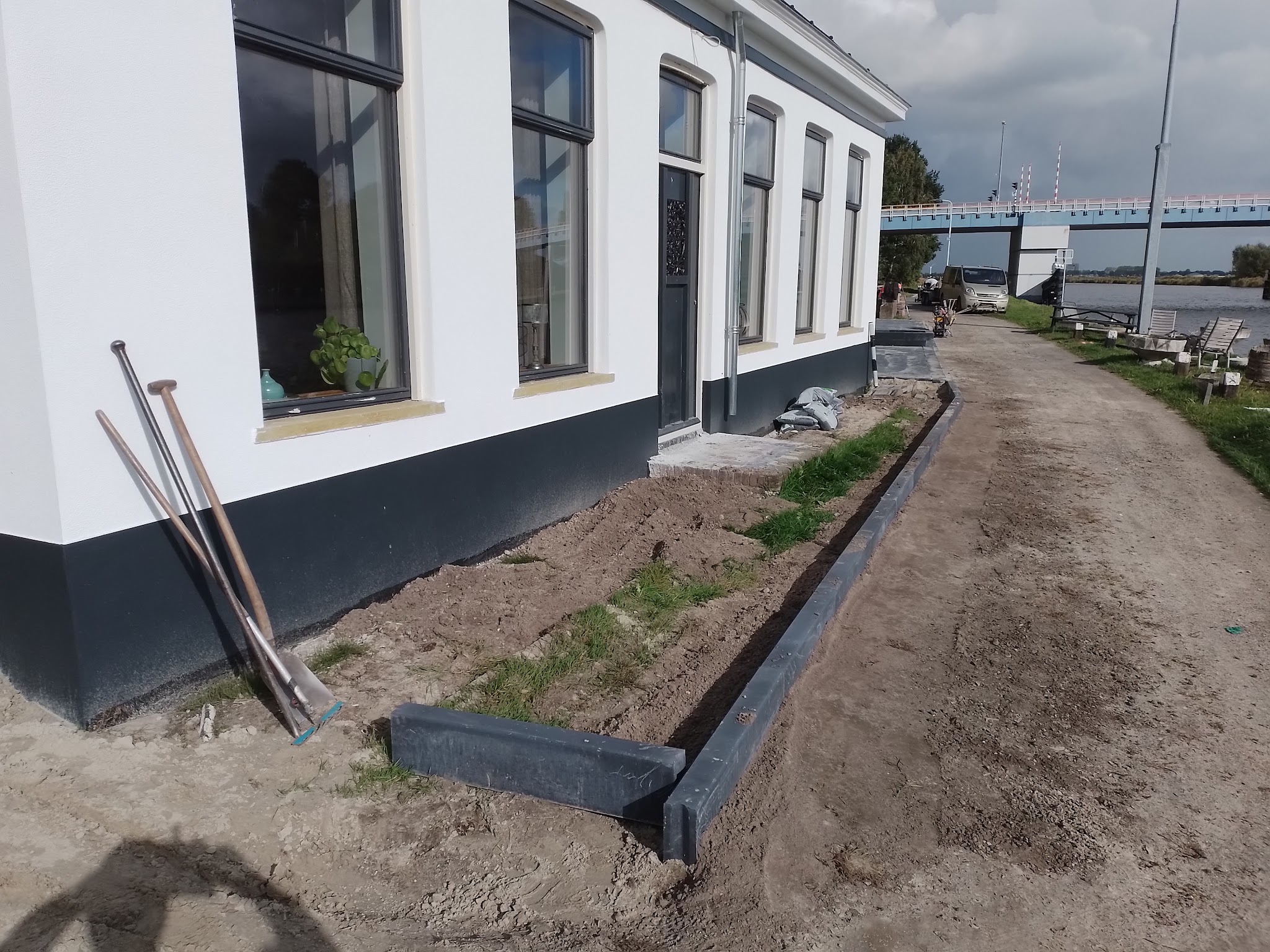 Zwarte opsluitbanden worden geplaatst langs een wit huis aan een vaart, met een ophaalbrug op de achtergrond