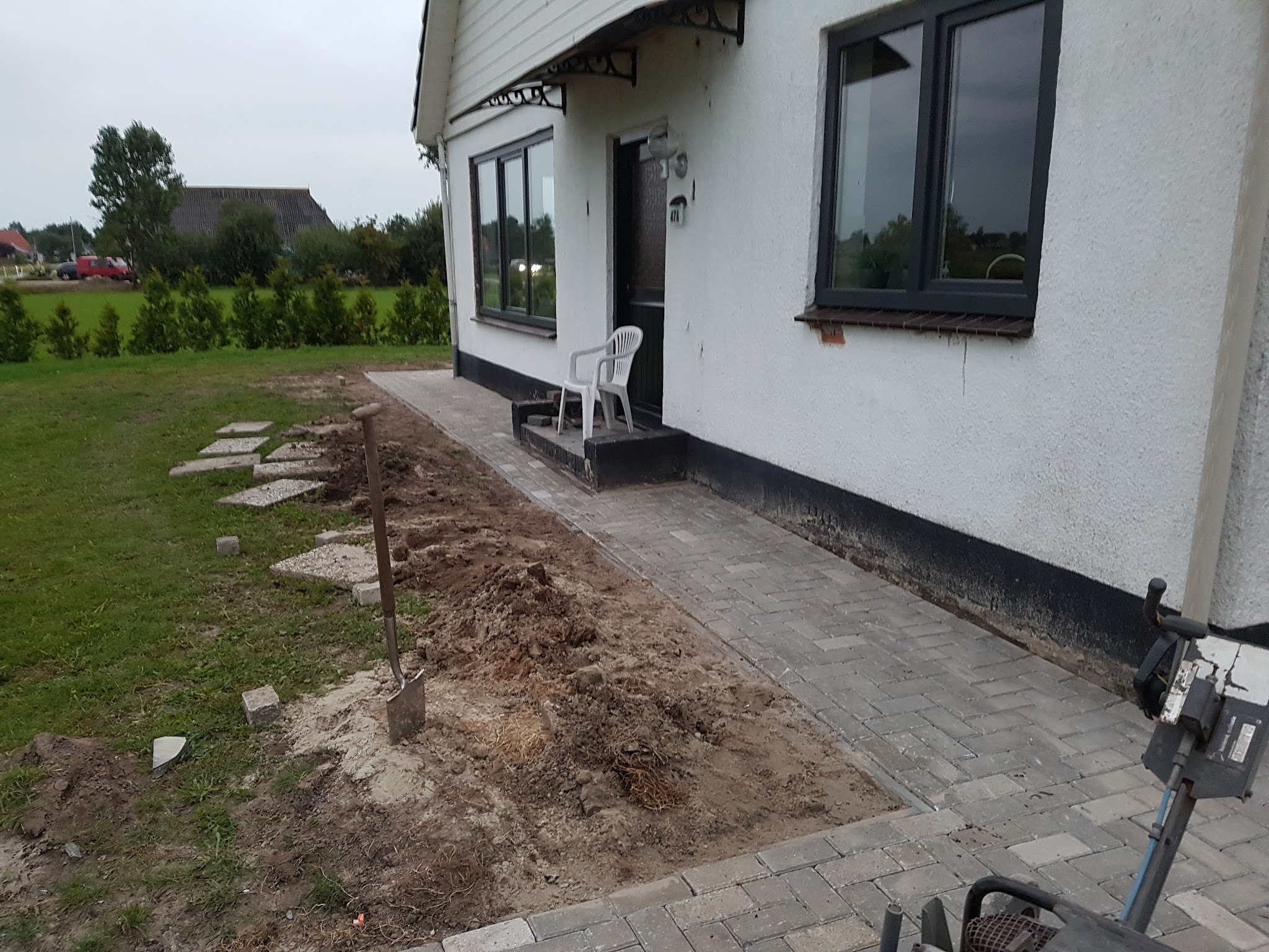 Grijs bestraat voorpad langs een wit huisje, stapstenen in het gras en een schop in verse aarde